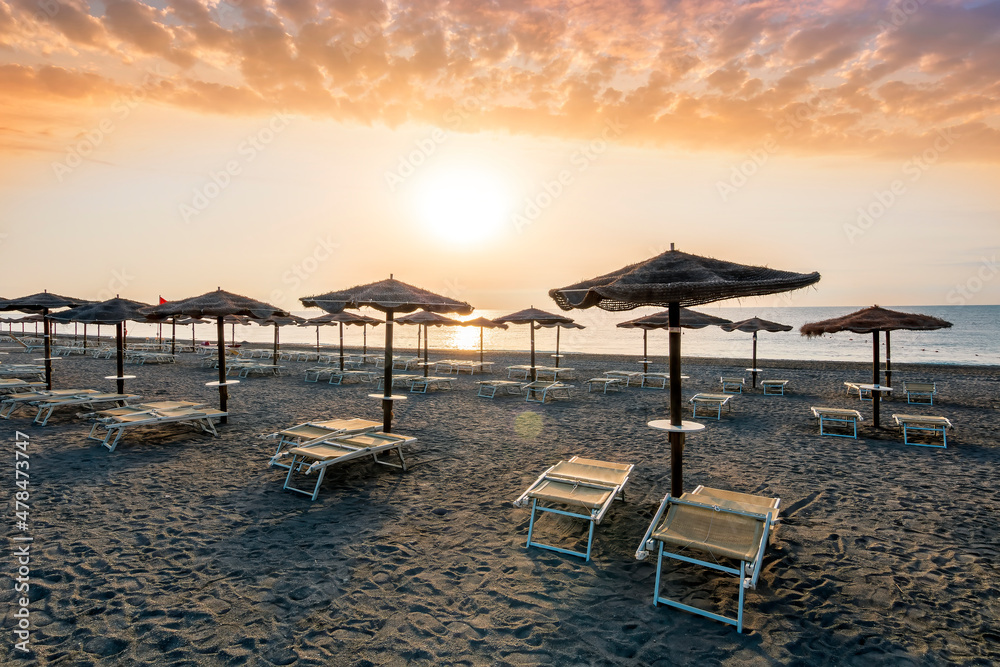 beautiful landscape of empty sunrise or sunset beach with chaise loungues and nice umbrellas with blue sea, sun glow and amazing cloudy sky on thr background