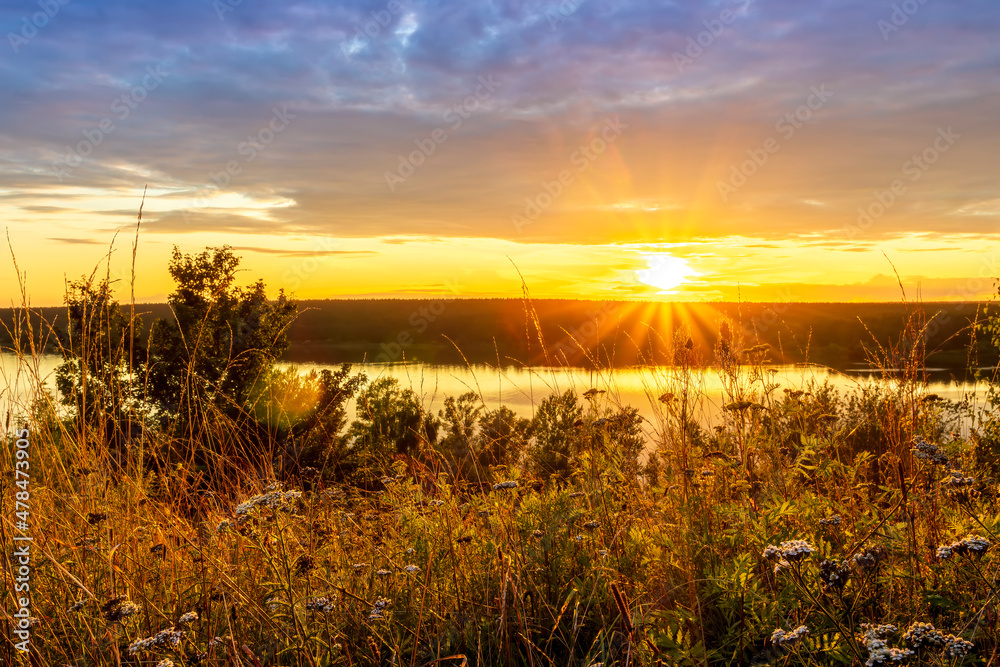 Amazing view at beautiful sunset or sunrise on a shiny lake from hill with green bushes, golden sun rays, calm water ,deep blue cloudy sky and forest on a background Amazing view at beautiful sunset or sunrise on a shiny lake from hill with green bushes, golden sun rays, calm water ,deep blue cloudy sky and forest on a background