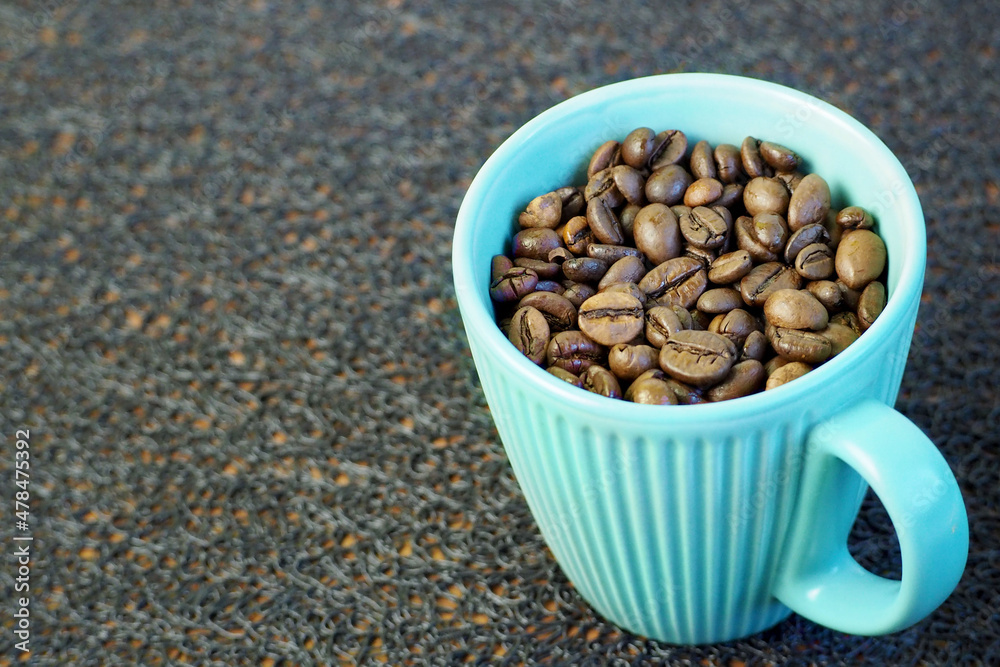 roasted dark brown coffee beans are poured into a turquoise mug with a handle on a black textured background