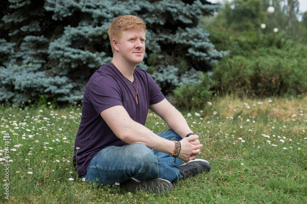 Portrait of redhead caucasian man wearing purple t-shirt and blue jeans and sitting on the grass in city park during lunch break at summer. Person looks away. Theme of rest in work break. Portrait of redhead caucasian man wearing purple t-shirt and blue jeans and sitting on the grass in city park during lunch break at summer. Person looks away. Theme of rest in work break.