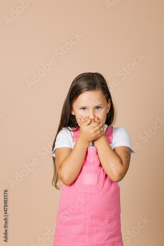 Portrait photo of female kid closing mouth with hands not talk with others with shining eyes and sullen look looking at camera wearing bright, pink jumpsuit and white t-shirt on beige background.