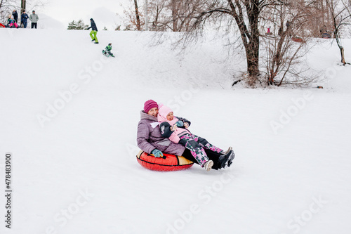 Photo of female Russian kid smiling and sliding on sled from hill with grandmother looking away wearing warm winter clothes in park. Astonishing background full of white color and snow. 