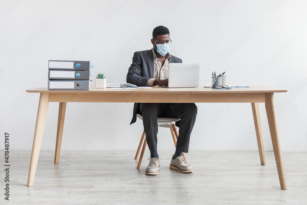 Young black businessman working from home office with laptop, wearing face mask during coronavirus quarantine