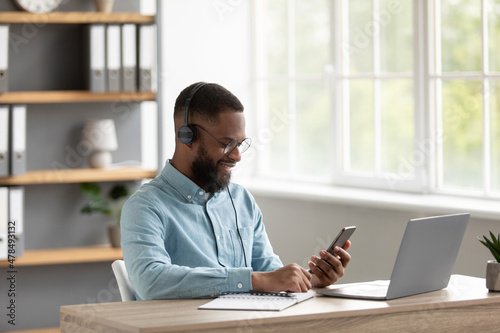 Satisfied attractive young black guy with beard in glasses and headphones chatting on phone at workplace with pc