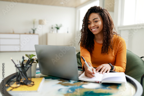 Black woman sitting at desk, using pc writing in notebook photo