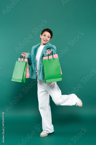 full length view of excited woman in faux fur jacket and white trousers posing with shopping bags on green © LIGHTFIELD STUDIOS