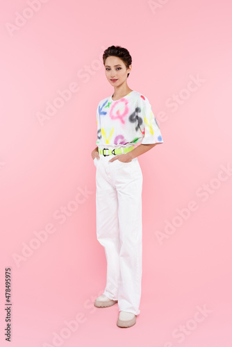 full length view of stylish smiling woman standing with hands in pockets of white trousers on pink