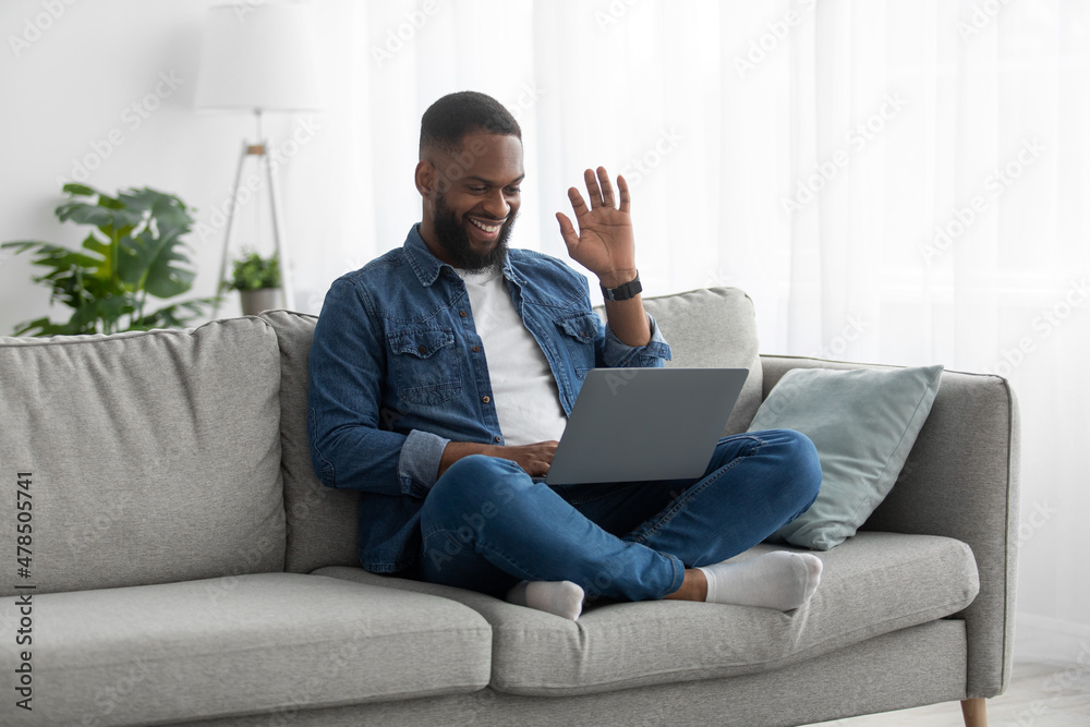 Happy attractive young african american male with beard sits on couch, greets, waves his hand to pc webcam Happy attractive young african american male with beard sits on couch, greets, waves his hand to pc webcam