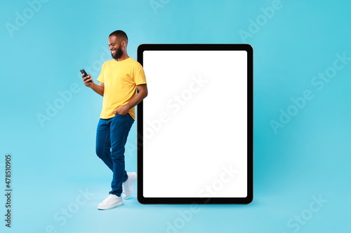 Optimistic young black man leaning on huge tablet computer with blank white screen, using mobile phone, mockup