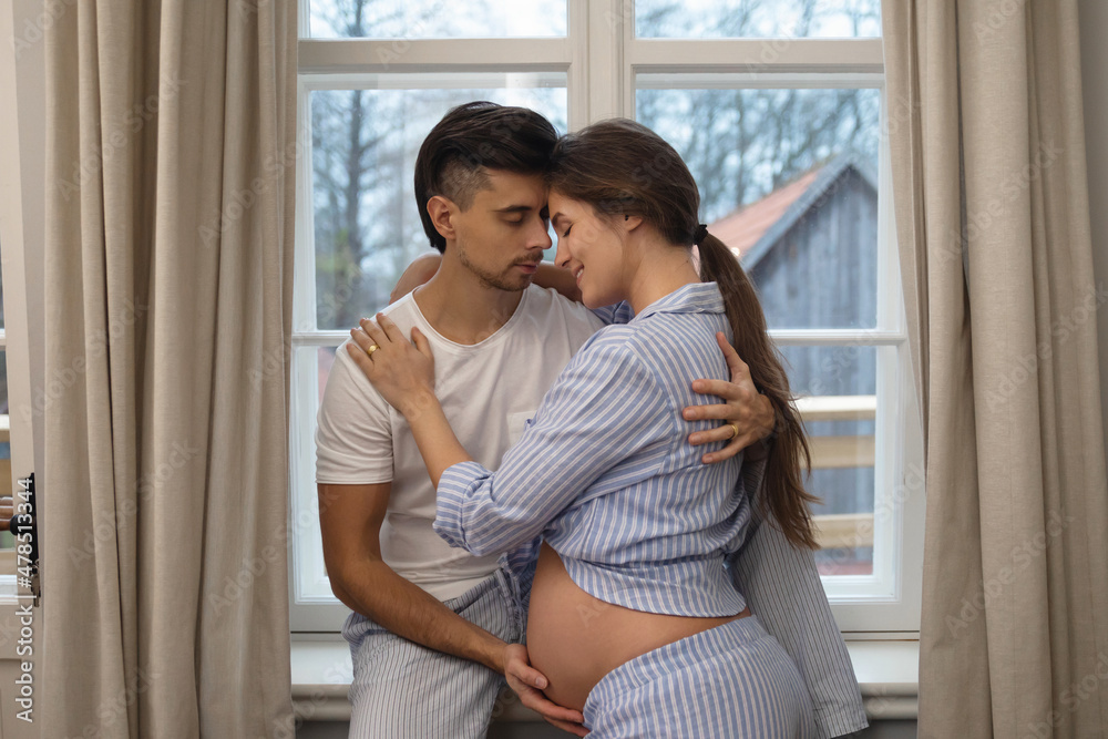 Naklejka premium Young sensual pregnant couple in embrace beside the window.