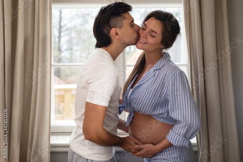Young sensual pregnant couple in embrace beside the window.