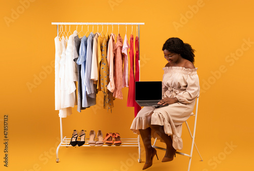 Beautiful young black woman sitting near clothes rack with laptop, offering mockup for web store design on screen