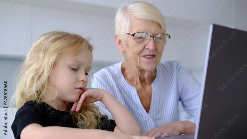 elderly caucasian woman grandmother and little curly granddaughter using laptop in the kitchen at home. online learning concept. woman with child spend time together elderly caucasian woman grandmother and little curly granddaughter using laptop in the kitchen at home. online learning concept. woman with child spend time together