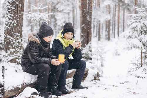 Happy teenage boys drinking tea from thermos and talking sitting together on log in winter snowy forest. Hot beverage in cold weather. Children having picnic in winter season outdoors. Local travel.