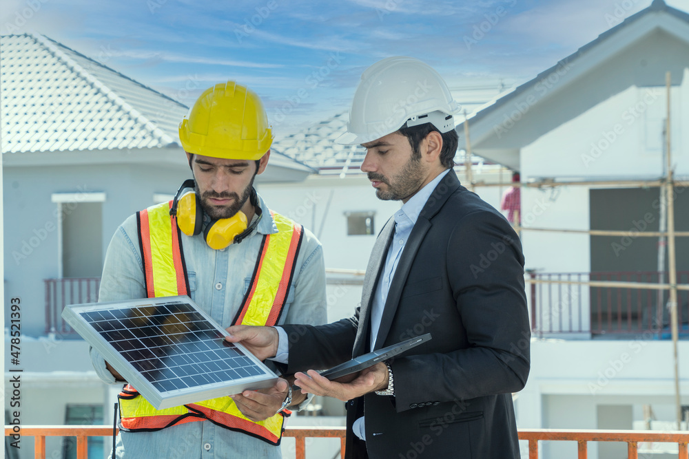 Foreman, an engineer holding a solar panel, consults with an architect to design a house, installing a solar system on a roof, standing at the construction site. Foreman, an engineer holding a solar panel, consults with an architect to design a house, installing a solar system on a roof, standing at the construction site.