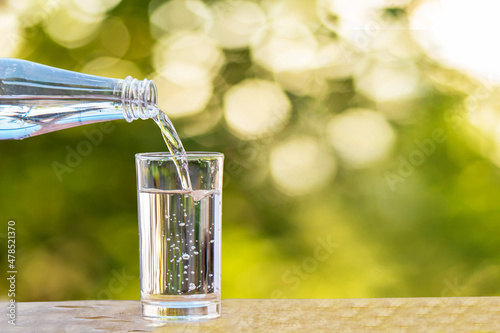 Pouring drinking water from bottle into glass on wooden tabletop on blurred fresh green nature background
