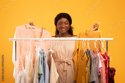 Shopping and fashion. Beautiful young black woman choosing outfits near clothing rack on orange studio background