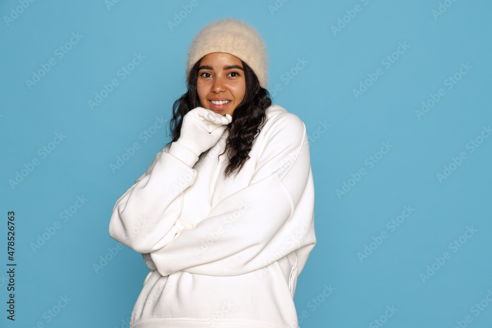 Smiling young beautiful dark skinned girl in warm white hoodie posing isolated on blue background.