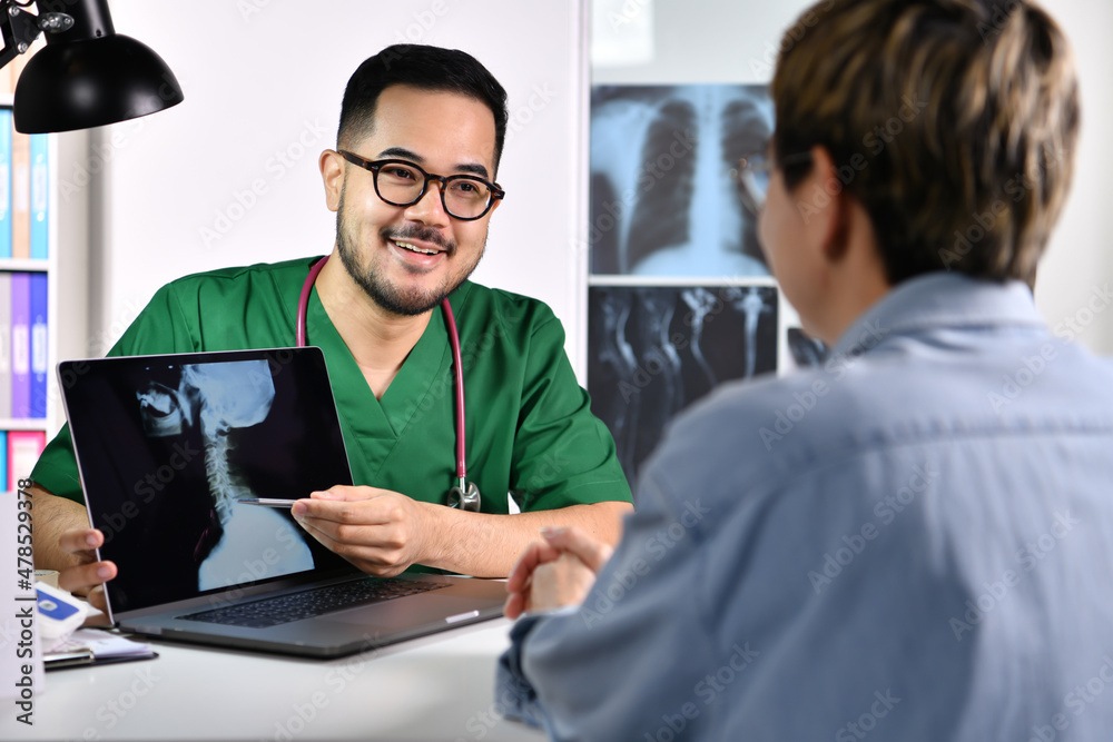 Obraz premium Kind comforting and attentive. Smiling young Asian male doctor in surgical uniform consulting and explaining female patient using laptop in examination room.