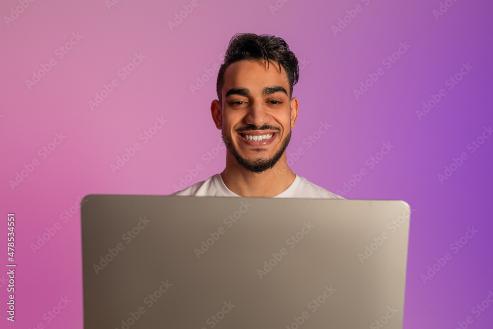 Handsome smiling young Arab man working on laptop, having video call, chatting online in neon light Handsome smiling young Arab man working on laptop, having video call, chatting online in neon light