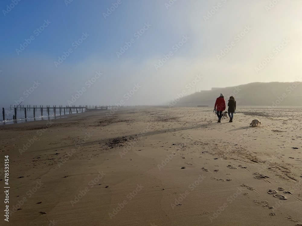 Beautiful landscape with female couple walking with dog on misty environment on sandy beach at Happisburgh in Norfolk coast in East Anglia uk with blue skies after fogbow on bright morning in December