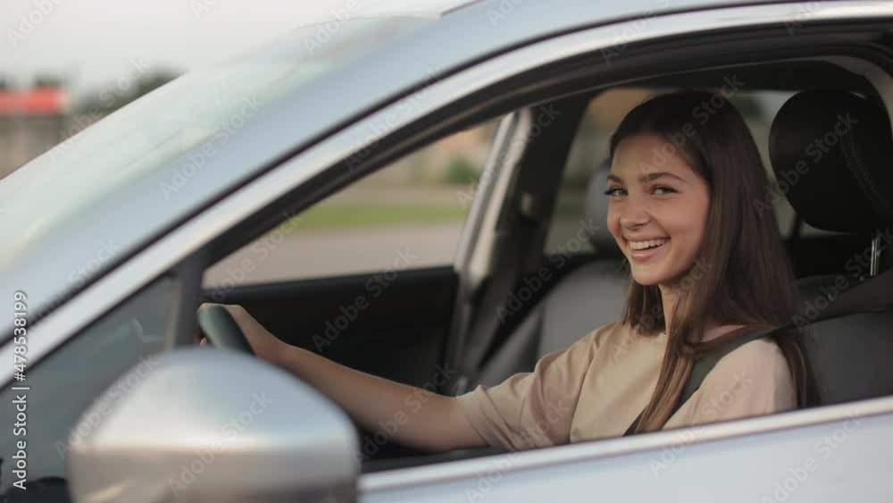A young woman is sitting in the car and smiling. She is turning her head and looking at the camera. She is keeping her hands on the steering wheel. The camera is moving around. 4K A young woman is sitting in the car and smiling. She is turning her head and looking at the camera. She is keeping her hands on the steering wheel. The camera is moving around. 4K