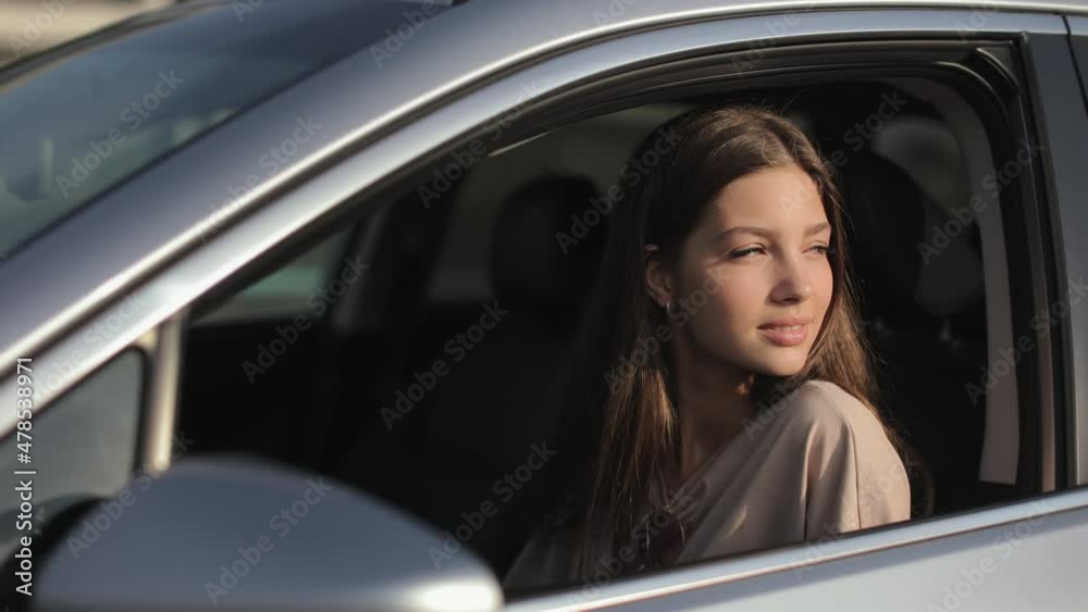 A young woman is sitting in the car in the driver's seat. She is turning her head to the side and starting laughing. The sun is shining on her. 4K