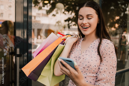 Shocked lady hold many bags and surfing internet with phone shopper woman dressed pink flower pattern dress carrying enjoying new clothes packs things after shopping buyings sales black friday concept