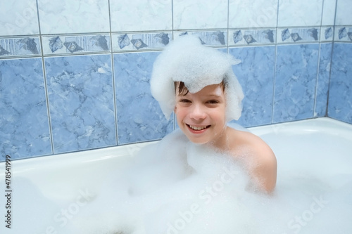 Portrait of happy child boy is playing with white foam sitting in bath at home  water games. Sensory games for children s development. He makes hat from foam on head. Looking at camera and smiling.