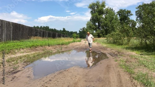 Caucasian senior man with walking stick moving through big puddle on the country road at summer season in Ukraine
