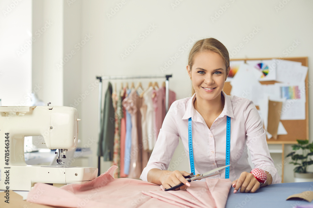 Fototapeta premium Portrait of smiling skillful young female seamstress at her workplace in sewing studio. Woman working with fabric and tailor's scissors looks at camera on background of her finished products.