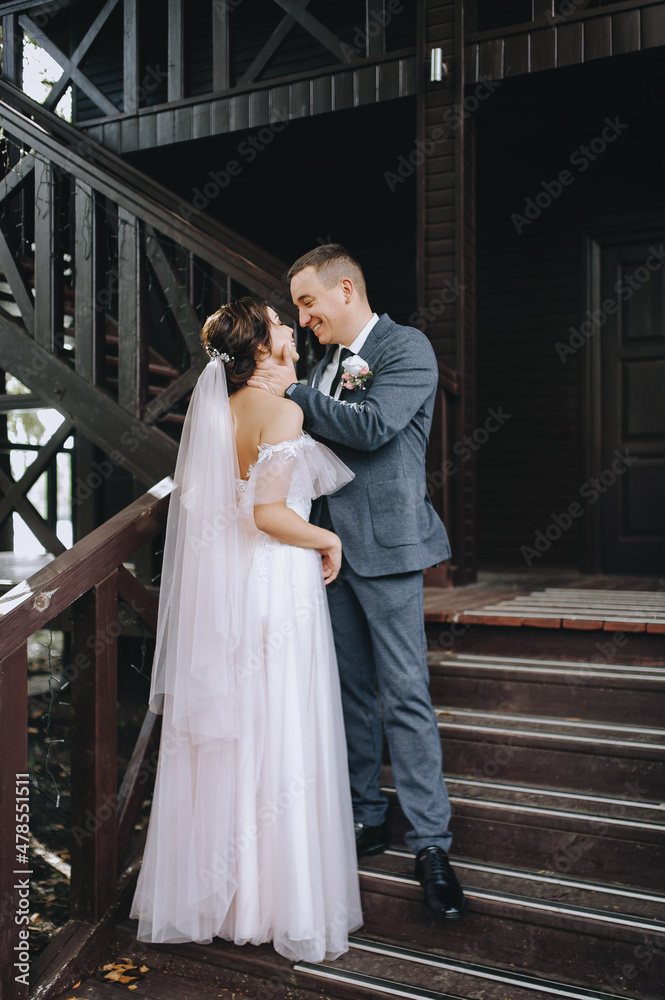 Fototapeta premium Stylish, young groom in a gray suit gently hugs a beautiful brunette bride in a white dress on the stairs, against the background of a wooden house. Wedding portrait of smiling newlyweds.
