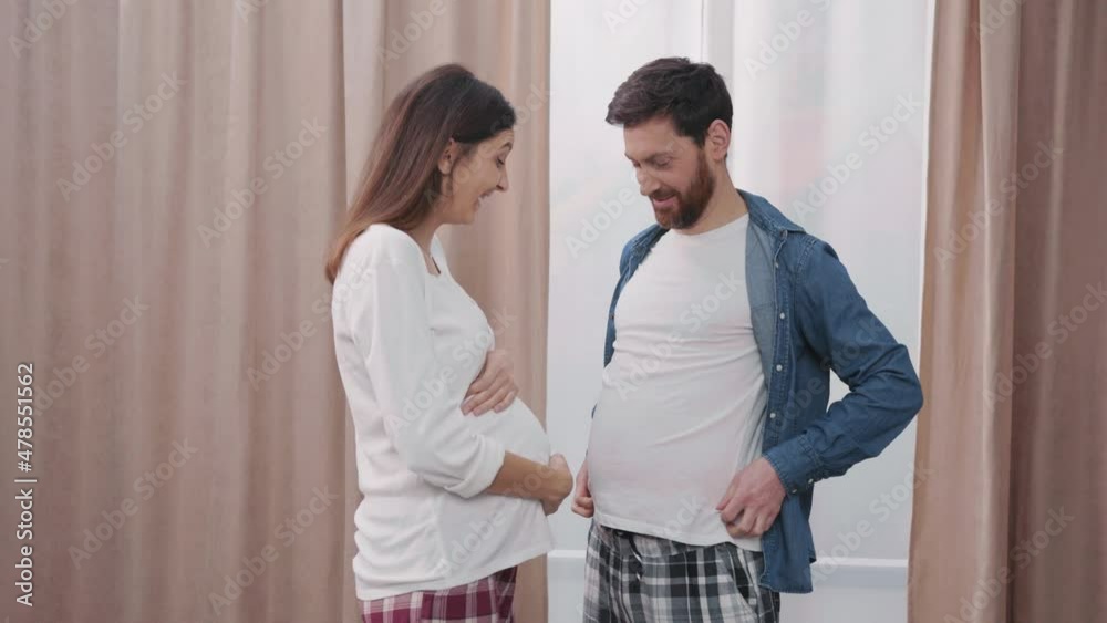 Lovely young woman expecting child laughing at her pregnant husband. Pretending funny man showing food tummy. Families fun. Lovely young woman expecting child laughing at her pregnant husband. Pretending funny man showing food tummy. Families fun.