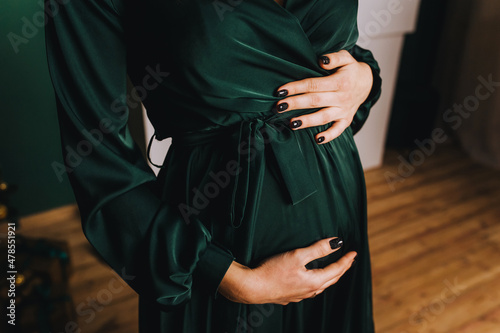 A pregnant girl in a green dress holds her big tummy with her hands. Portrait, photography.