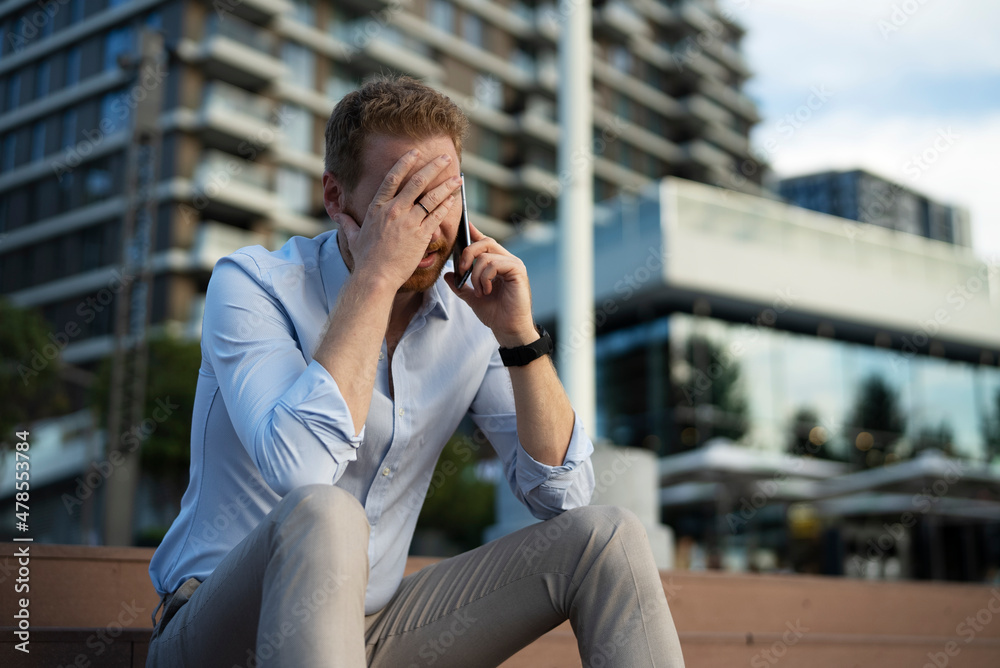 Businessman talking to the phone while sitting on the stairs. Angry upset man talking to the phone. Businessman talking to the phone while sitting on the stairs. Angry upset man talking to the phone.