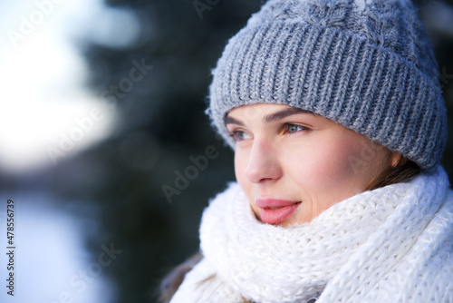 Close up beauty portrait of beautiful girl, pretty attractive woman in warm clothes knitted hat and white scarf is walking in winter snowy park at cold snow frosty day looking into distance. North