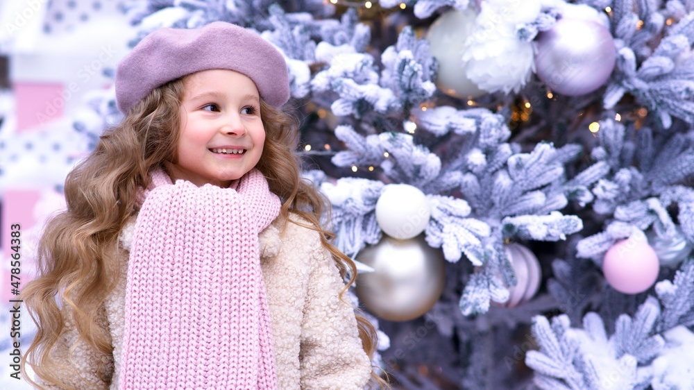 Portrait of cute happy positive cheerful kid girl adorable beautiful child is smiling outdoors in hat coat and scarf looking to right side. Christmas tree background. Banner, copy space place for text Portrait of cute happy positive cheerful kid girl adorable beautiful child is smiling outdoors in hat coat and scarf looking to right side. Christmas tree background. Banner, copy space place for text