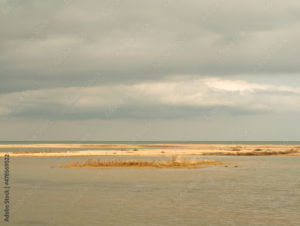 View of a shallow lagoon separated from the sea by a slanting sandy beach in early spring on a sunny day. Yellow shell-sand islands in seawater