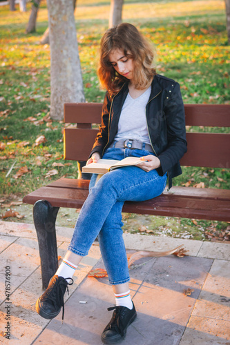 Cheerful teenager girl reading book on the bench at autumn park