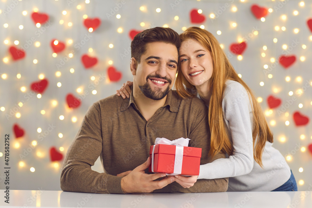 Saint Valentine's Day celebration, gift giving and romantic couple concept. Portrait of happy smiling young man and woman hugging and holding red present box against lights and hearts decor background Saint Valentine's Day celebration, gift giving and romantic couple concept. Portrait of happy smiling young man and woman hugging and holding red present box against lights and hearts decor background