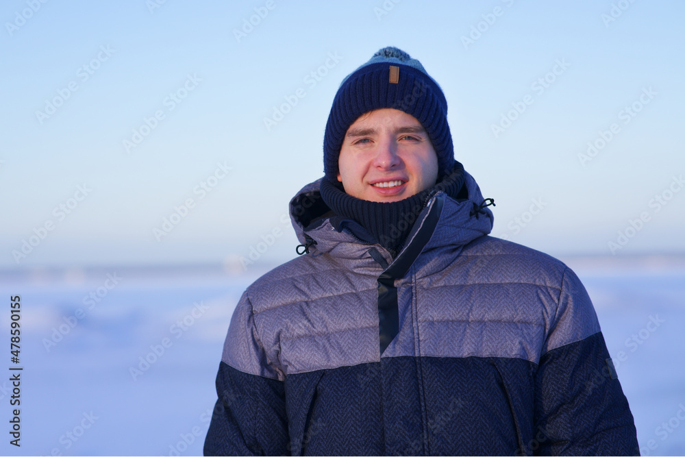 Portrait of happy cheerful positive guy, young handsome joyful man is smiling, enjoying walking outdoors in winter park at snowy frosty sunny day in warm clothes, hat, looking at camera. Wintertime.