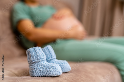 Little knitted socks for future child. Blue baby booties on sofa at the blur background of a pregnant woman resting on the sofa © Vadim