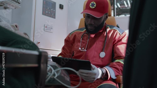 African American doctor in red uniform checking vital signs monitor of patient and entering medical data into tablet while riding emergency medicine vehicle to hospital photo