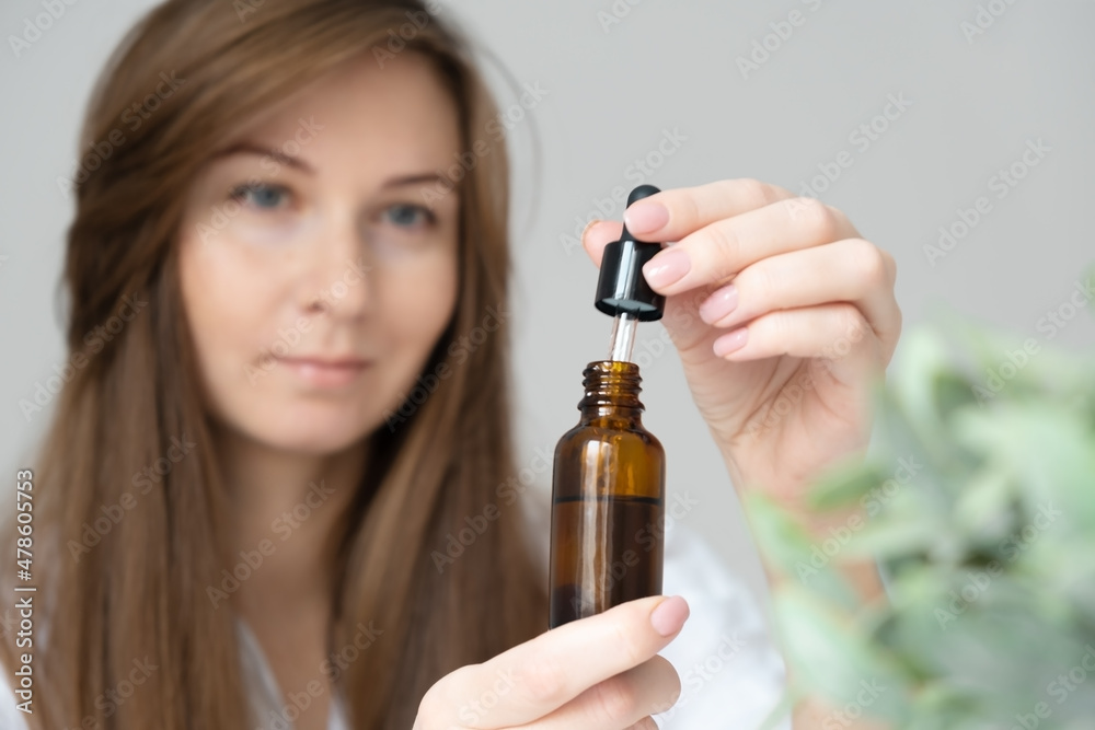 Young caucasian long haired brunette woman defocused doing body,skin care beauty procedures at home,holding glass bottle with serum,oil,bio natural cosmetics for hair or face.Selective focus Young caucasian long haired brunette woman defocused doing body,skin care beauty procedures at home,holding glass bottle with serum,oil,bio natural cosmetics for hair or face.Selective focus