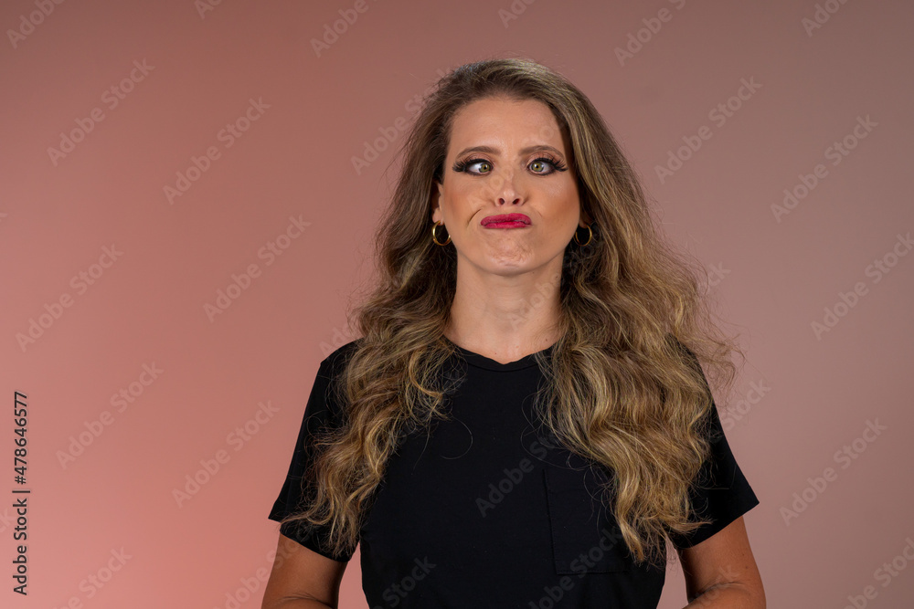 Retrato de mulher loira de olhos verdes, cabelo cacheado, roupa preta em fundo cor salmão no estúdio. Jovem olhando para a câmera e fazendo careta de brincadeira. Retrato de mulher loira de olhos verdes, cabelo cacheado, roupa preta em fundo cor salmão no estúdio. Jovem olhando para a câmera e fazendo careta de brincadeira.