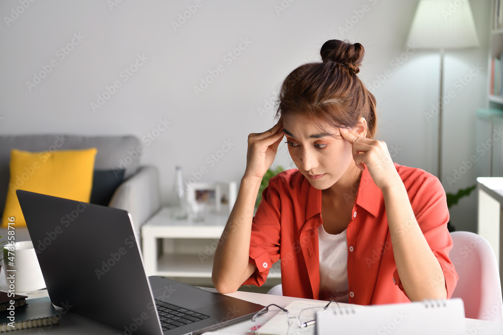 Young Asian woman sitting looking exhausted and tired during working at home using laptop. Overworked tired Asian female student worker massaging eyes and temples from fatigue after computer work. Young Asian woman sitting looking exhausted and tired during working at home using laptop. Overworked tired Asian female student worker massaging eyes and temples from fatigue after computer work.