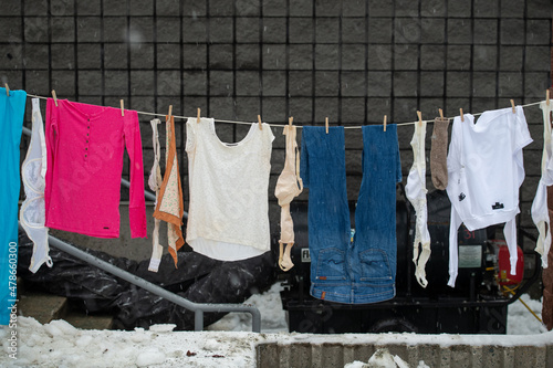 A colorful line of clothes hanging outside air drying with a brick building in the background. The line has a number of bras, shirts, jeans, and tops. The cloth brassieres are of large sizes.