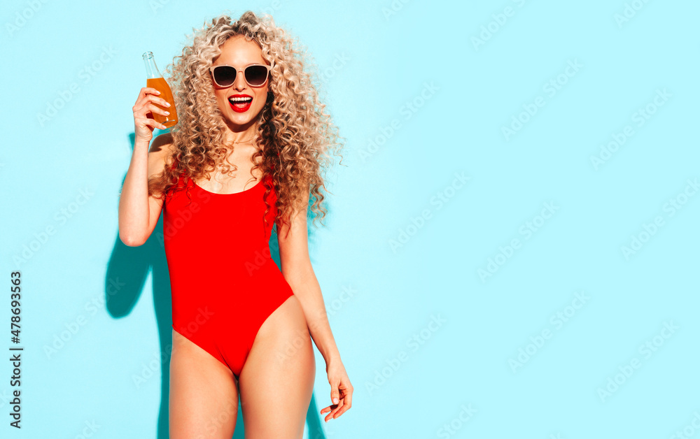 Young beautiful smiling woman posing near blue wall in studio.Sexy model in red swimwear bathing suit.Positive female with afro curls hairstyle.Drinks lemonade from a glass bottle.Happy and cheerful Young beautiful smiling woman posing near blue wall in studio.Sexy model in red swimwear bathing suit.Positive female with afro curls hairstyle.Drinks lemonade from a glass bottle.Happy and cheerful