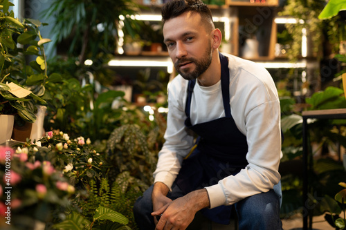 a satisfied male florist in a blue apron with a pruner in his hands sits with a smile looking at the camera © Ivan Traimak
