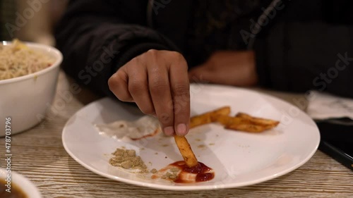 Close up view of guy sitting at table in the restaurant and eating French fries with mayonnaise and tomato sauce from a white plate. Guy wearing black jacket. photo